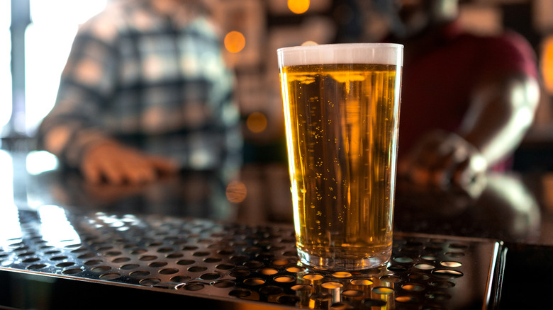 a freshly poured pint of lager on a bar with two people blurred in the background