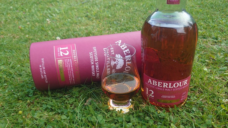 A filled tasting glass of Aberlour 12 shown on grass with the bottle next to it and the packing behind on its side