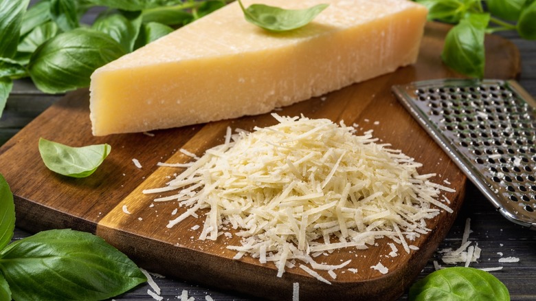 Grated hard cheese on a wooden board next to a cheese grater, and cheese wedge, and some fresh basil