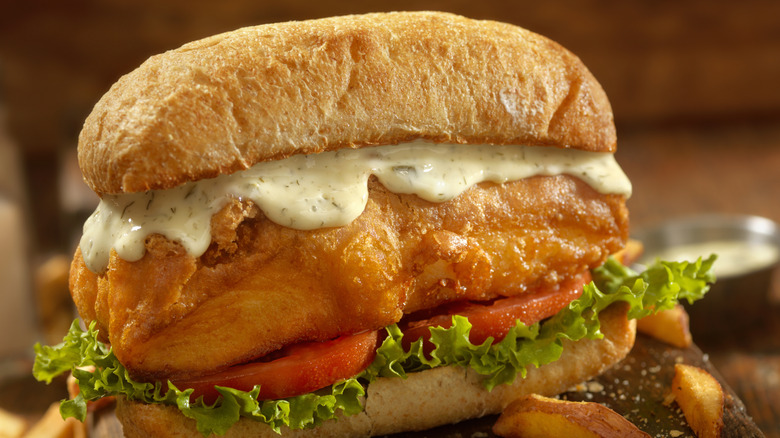 A fried fish sandwich dressed with lettuce, tomato, and tartar sauce, with a few French fries scattered around it