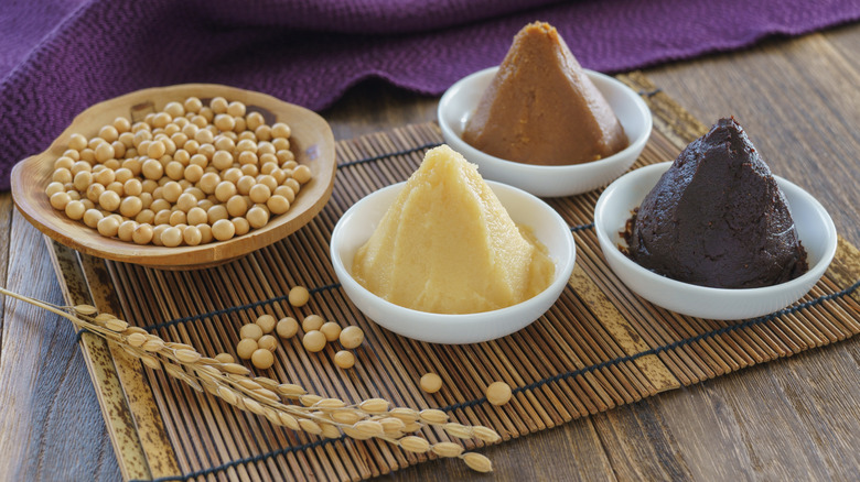 Bowls of white, red, and hatcho miso next to soybeans and stem of wheat