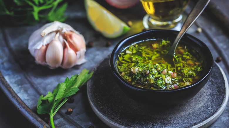 chimichurri in bowl with a spoon, surrounded by fresh herbs and garlic
