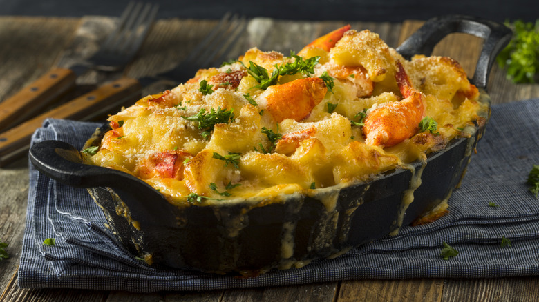 a cast iron dish filled with baked lobster mac and cheese topped with fresh herbs sitting atop a folded napkin on a wooden table