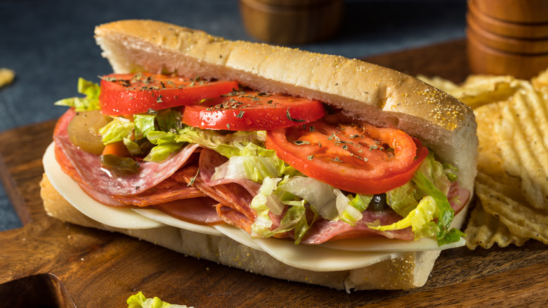 Italian sub sandwich with tomatoes, lettuce, cold cuts, and cheese sitting on board next to crinkle-cut potato chips