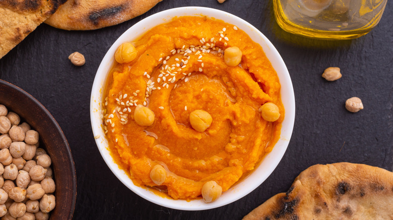 A bowl of orange-colored pumpkin hummus, surrounded by a bottle of olive oil, several pieces of pitta bread, and a bowl of chickpeas, with several chickpeas scattered on the table around them