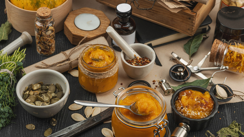 A work surface with several containers of pumpkin purée, along with a bowl of pumpkin seeds, a dark bottle, several glass jars of dry seasoning, a mortal and pestle, a pair of scissors, and a bundle of fresh herbs