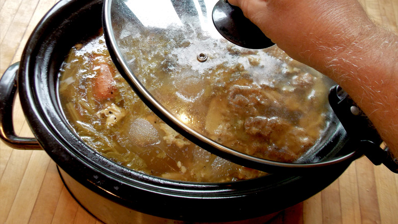 A hand removing the lid from a crock pot revealing an amber colored soup with bits of meat and vegetables floating in it