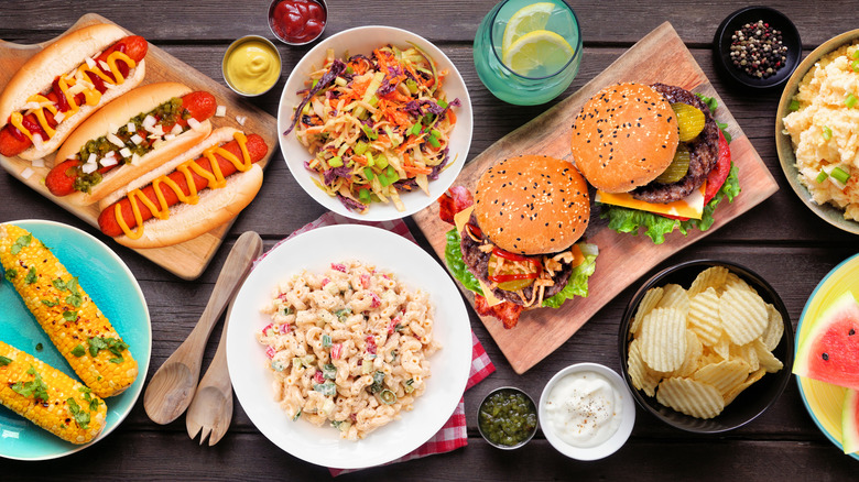 Summer cookout dishes including burgers, hot dogs, coleslaw, macaroni salad, potato chips, and corn on the cob on dark wood table