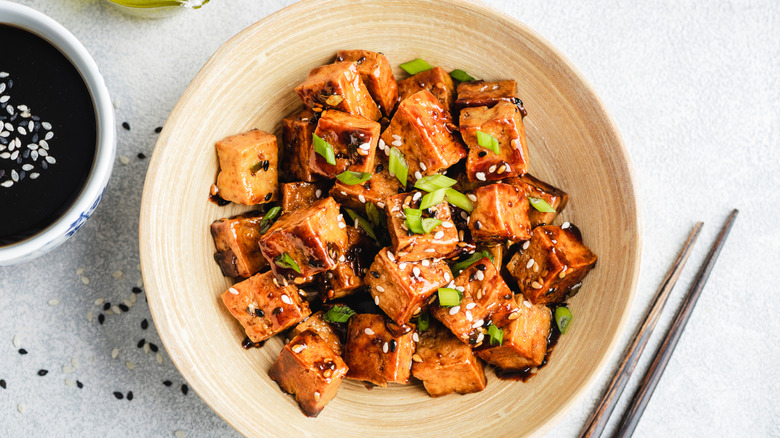 Tofu covered in sauce in a bowl with sesame seeds and green onions with extra sauce and chopsticks on the side