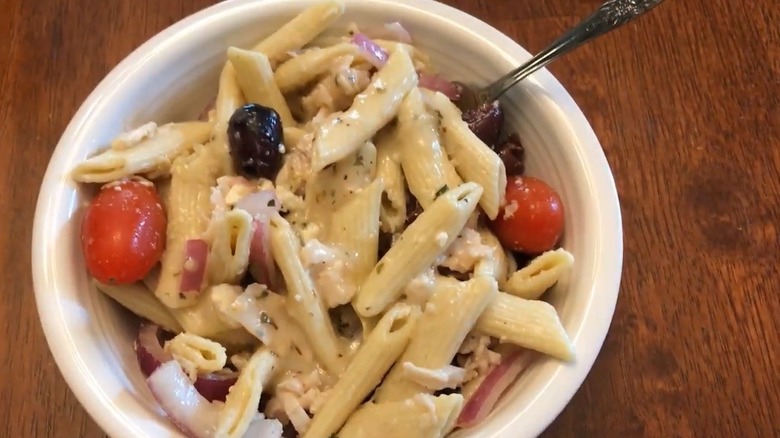 A white bowl full of penne pasta, kalamata olives, cherry tomatoes, red onion, and feta crumbles with dressing