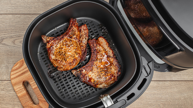 Two browned beef steaks inside air fryer basket next to black air fryer on wooden surface