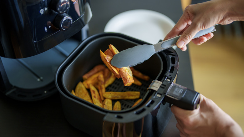 Person removing potato wedges from an air fryer basket using tongs