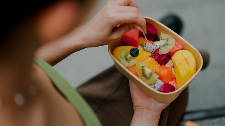 Person eating container of assorted fruit with kiwi, dragonfruit, blueberries, oranges, and others.