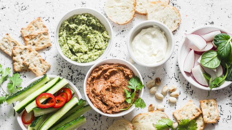 Different dips in white bowls on top of a countertop with bread, crackers, and crudite surrounding it.