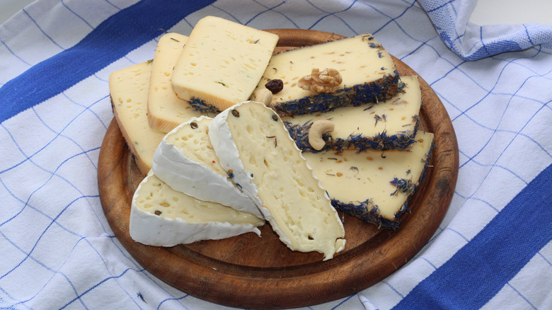 A round wooden platter of soft cheeses on top of a blue and white windowpane napkin.