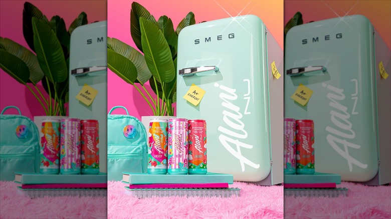 Cans of Alani Nu energy drinks stacked on notebooks next to a backpack and Smeg mini fridge