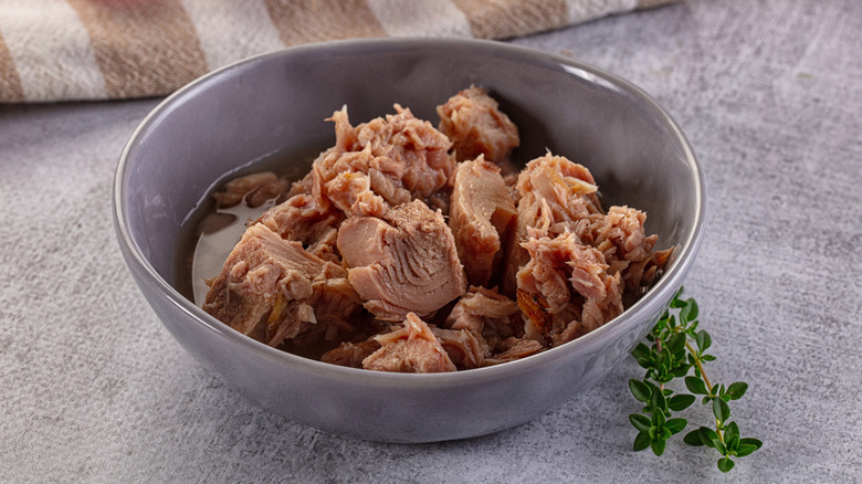 A bowl of canned tuna filet sitting on a blue tablecloth with a brown and white striped cloth in the background and a sprig of parsley in the foreground