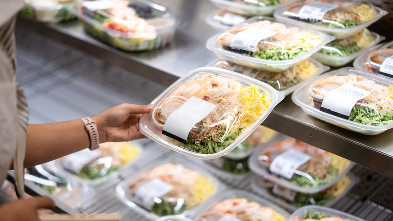 A person holding a grocery store prepared meal in their hands with more prepackaged meals in the background.