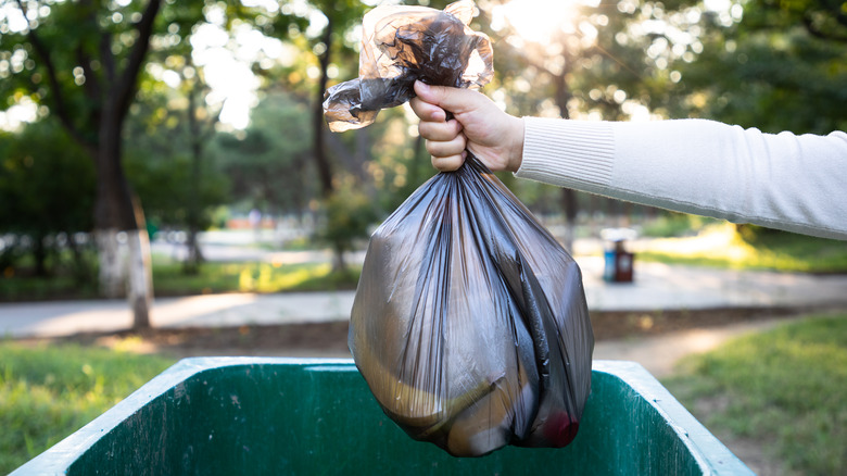 A person putting a black garbage bag into a green outdoor trash can