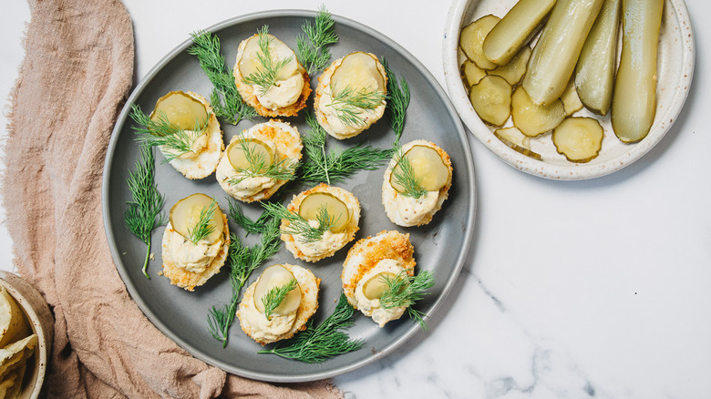 Overhead view of pickle and dill topped deviled eggs on a grey plate, surrounded by pickles, beer, and dill pickle chips.