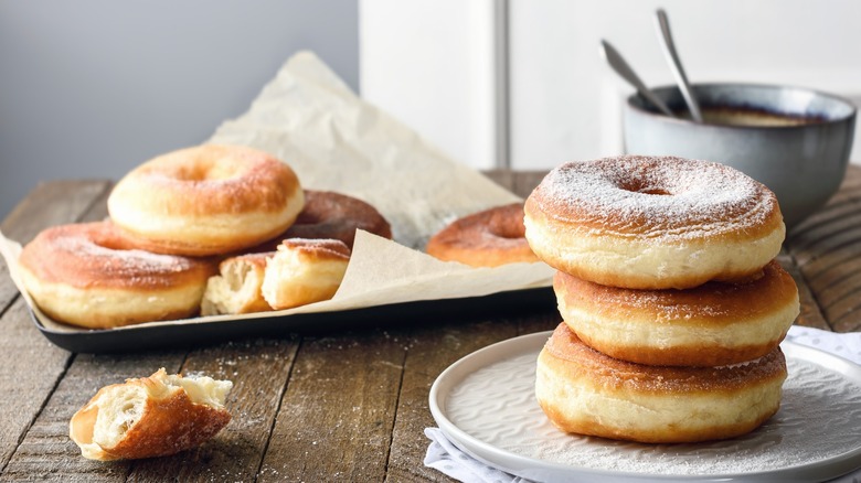 Homemade donuts stacked on a plate and more on a different plate behind dusted with powdered sugar