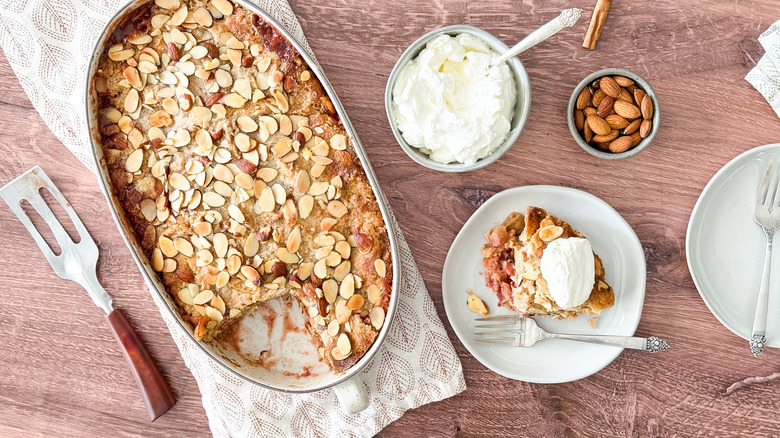 Almond-rhubarb from scratch dump cake in serving dish with almond whipped cream, plates, and almonds on table