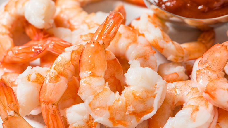 a close-up photo of a plate piled with cooked shrimp with a small metal dish of cocktail sauce in the background