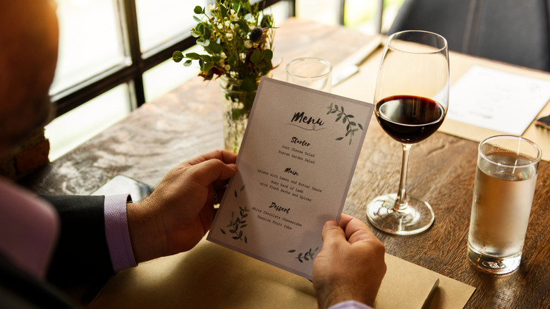 A man holding a restaurant menu up with a glass of red wine and some greenery in a vase on the table