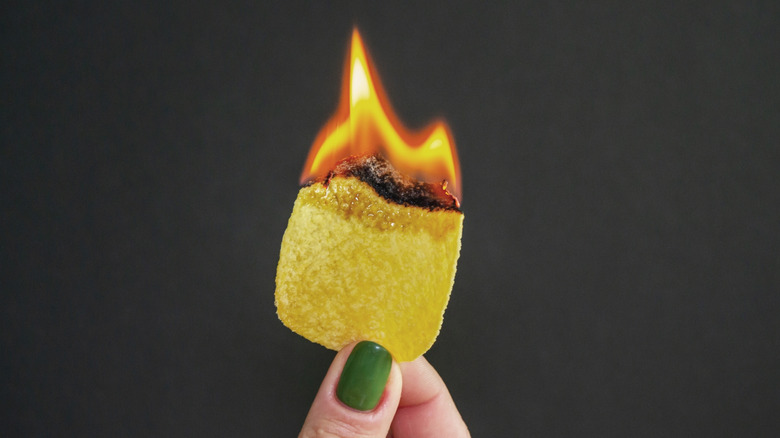 A hand holds a lit potato chip burning with a bright flame.