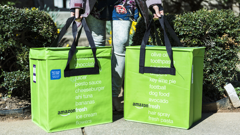 Two green Amazon Fresh bags being held by a person in front of a bush