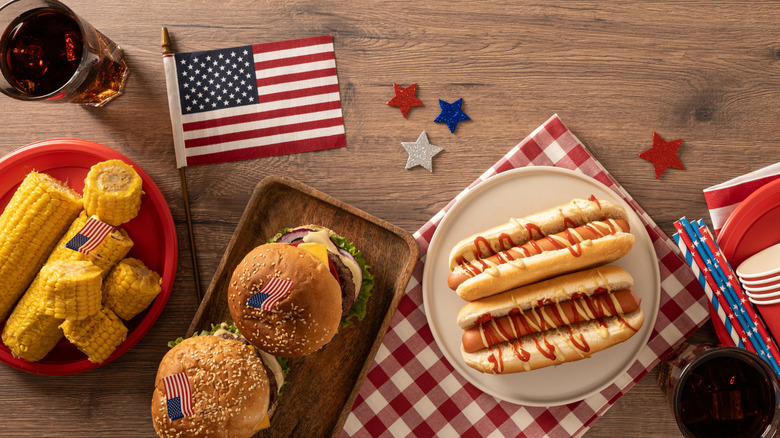Assorted cookout food, including hamburgers, hotdogs, and corn on the cob, sitting on a wooden table with a glass of soda and various patriotic U.S. symbols