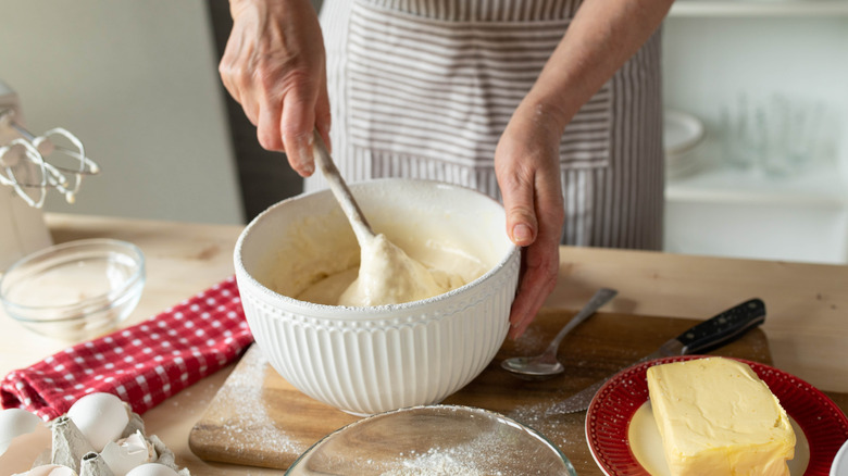 A person mixing dough in a white bowl, surrounded by ingredients like eggs and butter.