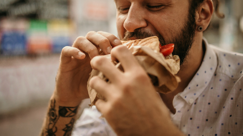 man with wrist tattoos bites into generic sandwich