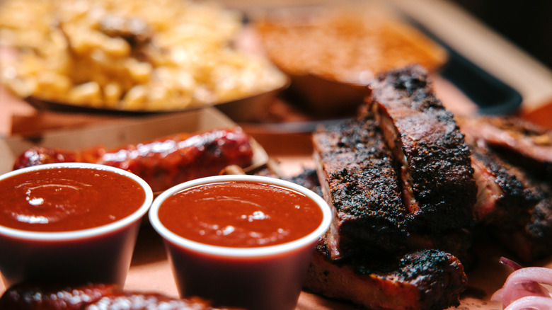 Close-up of BBQ ribs, sausage, and blurred sides with cups of sauce