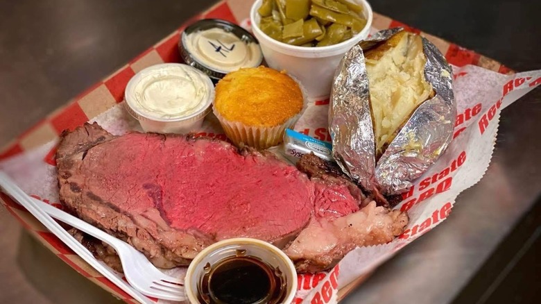 Smoked prime rib on a tray with a baked potato, beans, sauces, and cornbread