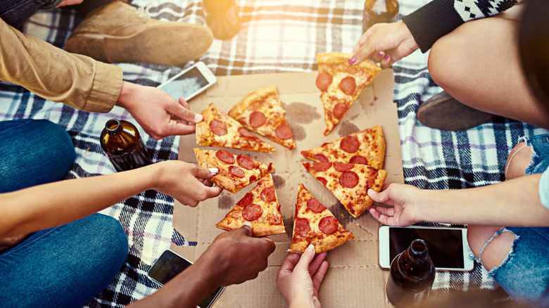 People eating pizza at outdoor picnic out of a cardboard box on a plaid blanket with brown glass bottles and phones.