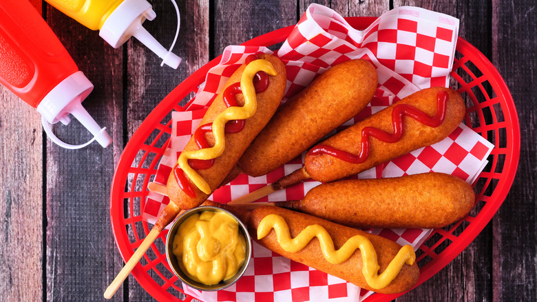 Five corn dogs in a red plastic basket lined with red and white checkered paper and drizzled with mustard and ketchup