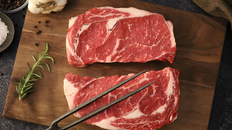 Two raw beef steaks sit on a wooden cutting board with thyme, garlic, peppercorns, and a carving fork.