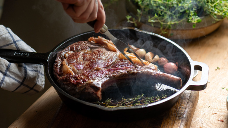 A person uses a carving fork to hold a tomahawk steak in a hot cast iron skillet with herbs and pearl onions.