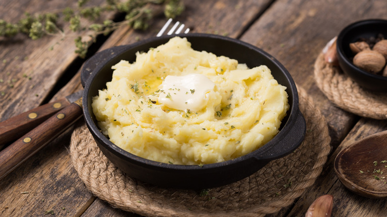A cast iron bowl of mashed potatoes with butter and some dry parsley on top, sitting on a woven trivet on a wooden table