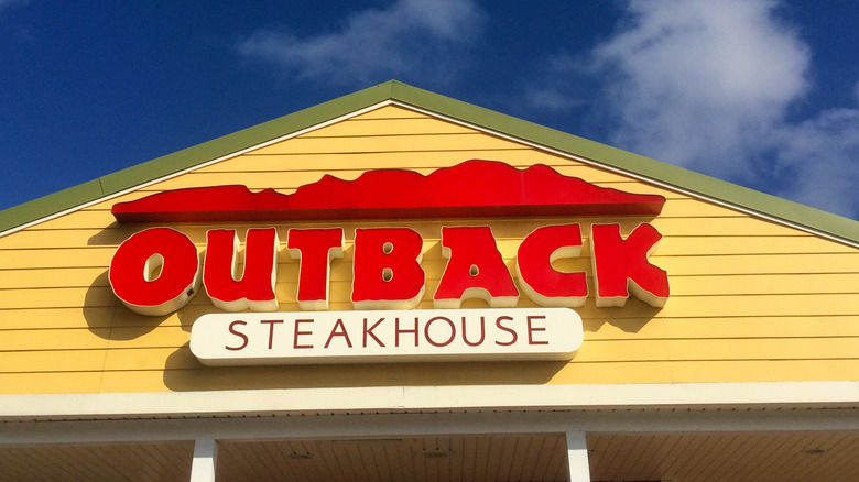 A red Outback Steakhouse sign on a peaked yellow roof with a blue sky behind