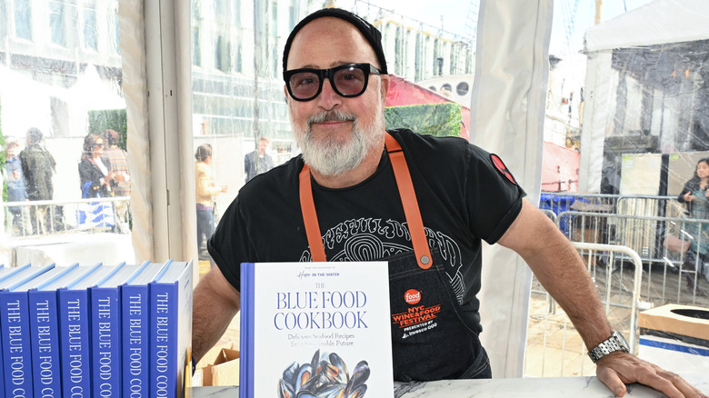 Andrew Zimmern smiles behind his cookbook wearing an apron