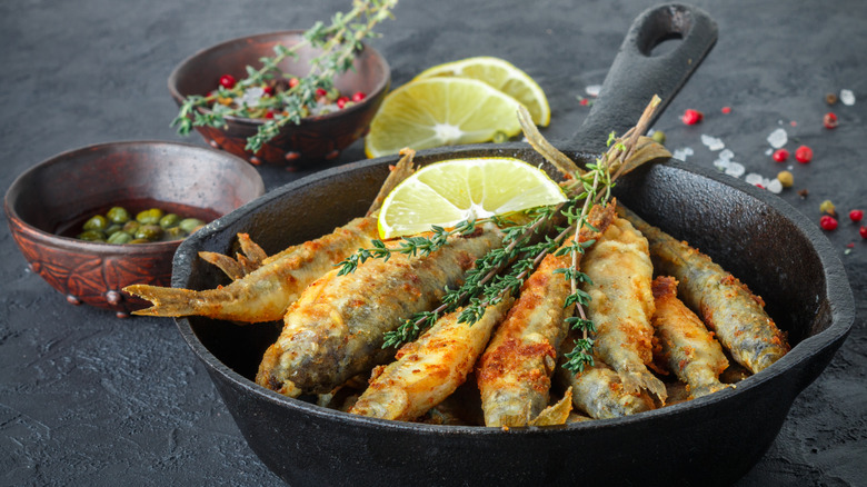 A cast iron pan of fried smelts next to bowls filled with capers and seasonings