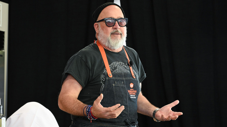 Andrew Zimmern wearing a denim apron and addressing the audience during a culinary demonstration