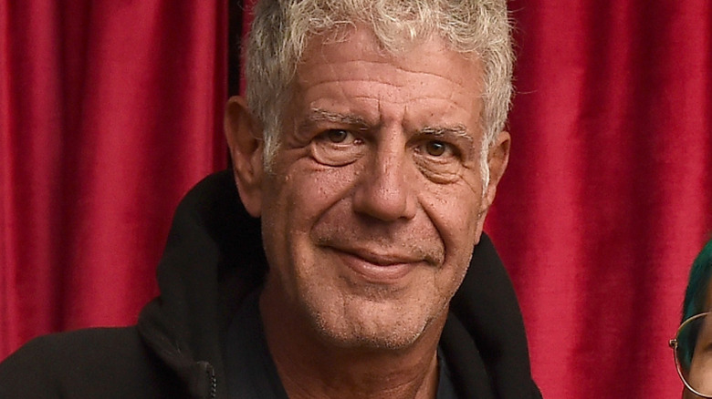 Close on Anthony Bourdain's face, he is wearing a black hoodie, and there is a red curtain behind