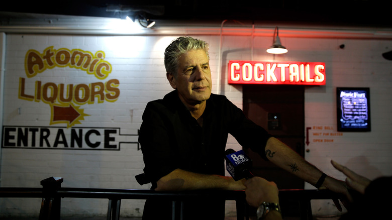 Anthony Bourdain outside Atomic Liquors, with a neon "COCKTAILS" sign and a microphone.