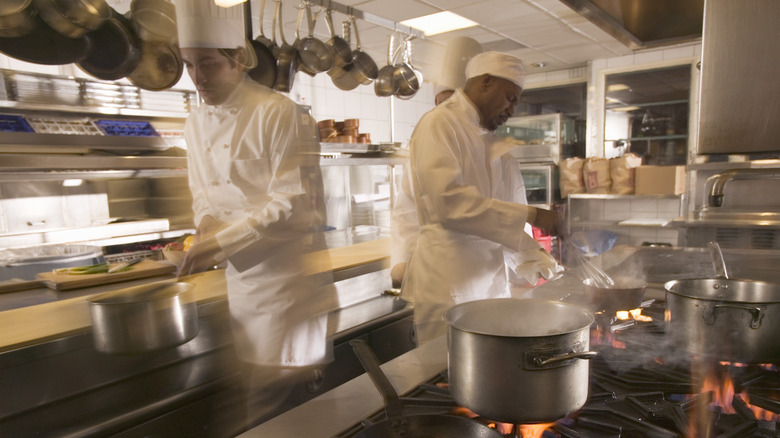 Two chefs dressed in traditional white uniforms working in a bustling restaurant kitchen, with several steel pots steaming on the stove in front of them.