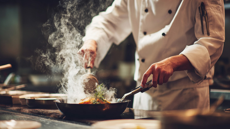A person in a chef jacket in a commercial kitchen making something in a skillet