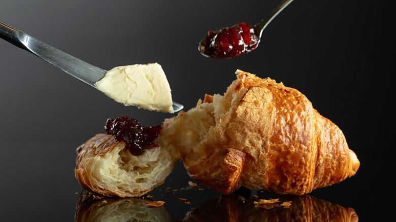 A croissant with an end torn off and a knife hovering above with butter and a spoon hovering with jam