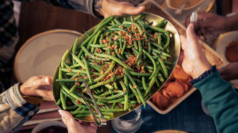 Two people passing a dish of green beans topped with fried onions